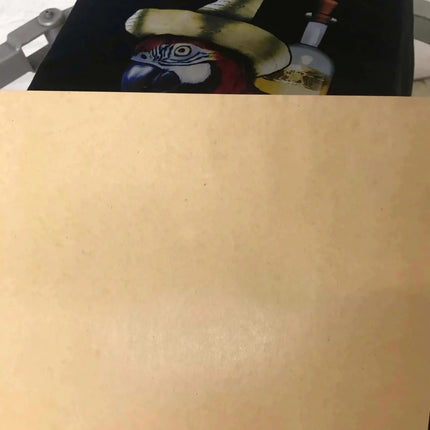 Kraft Dark Garment Pressing Paper 16" x 20" on dark fabric, bold red letters above a yellow hat and blue parrot graphic.